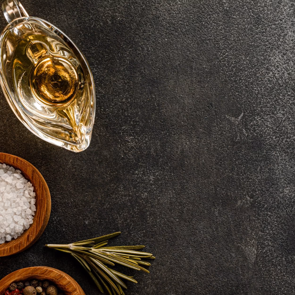 a glass jug of oil beside a wooden bowl of rock salt and a sprig of rosemary on dark stone