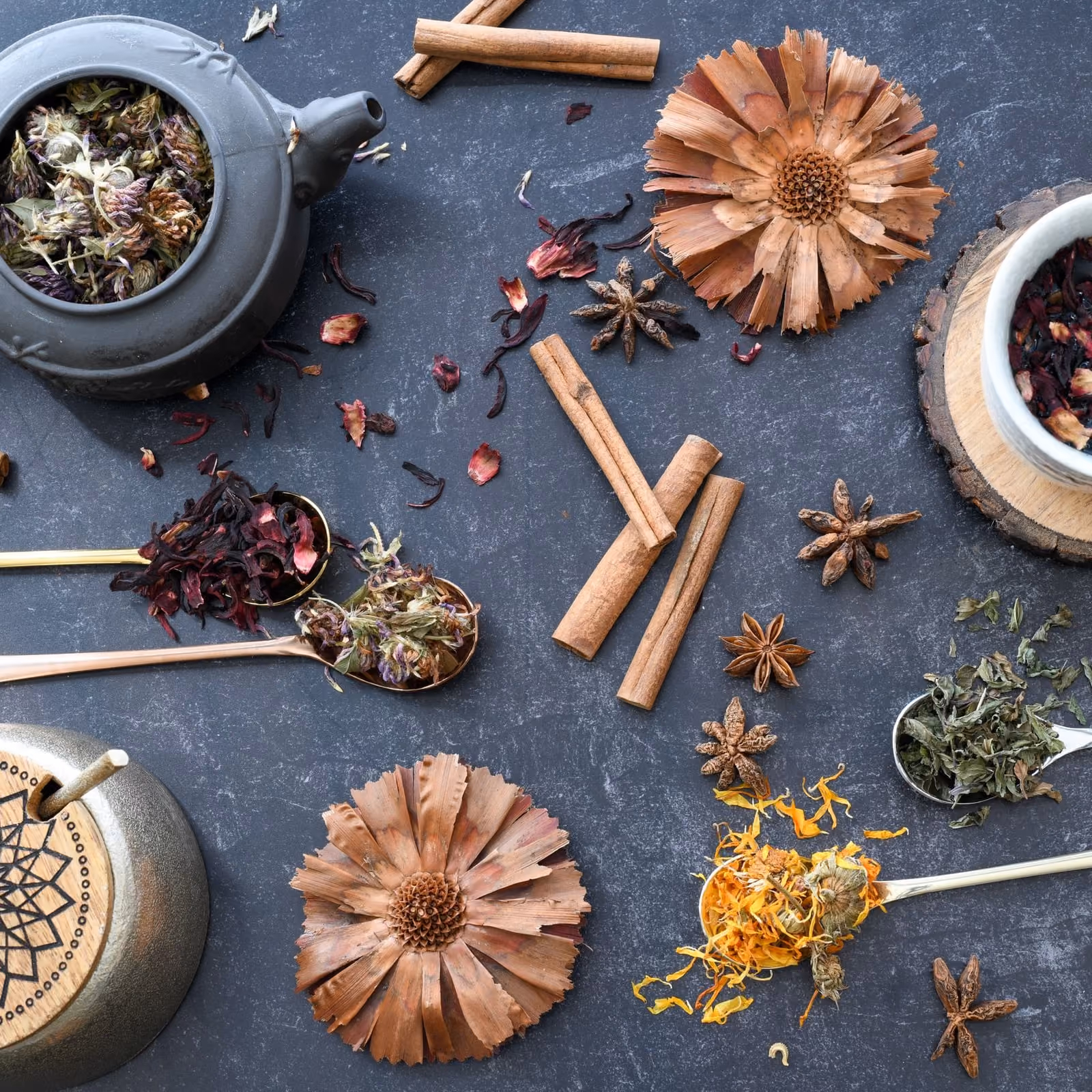 dried herbs, hibiscus, cinnamon and star anise on brass spoons across a slate surface