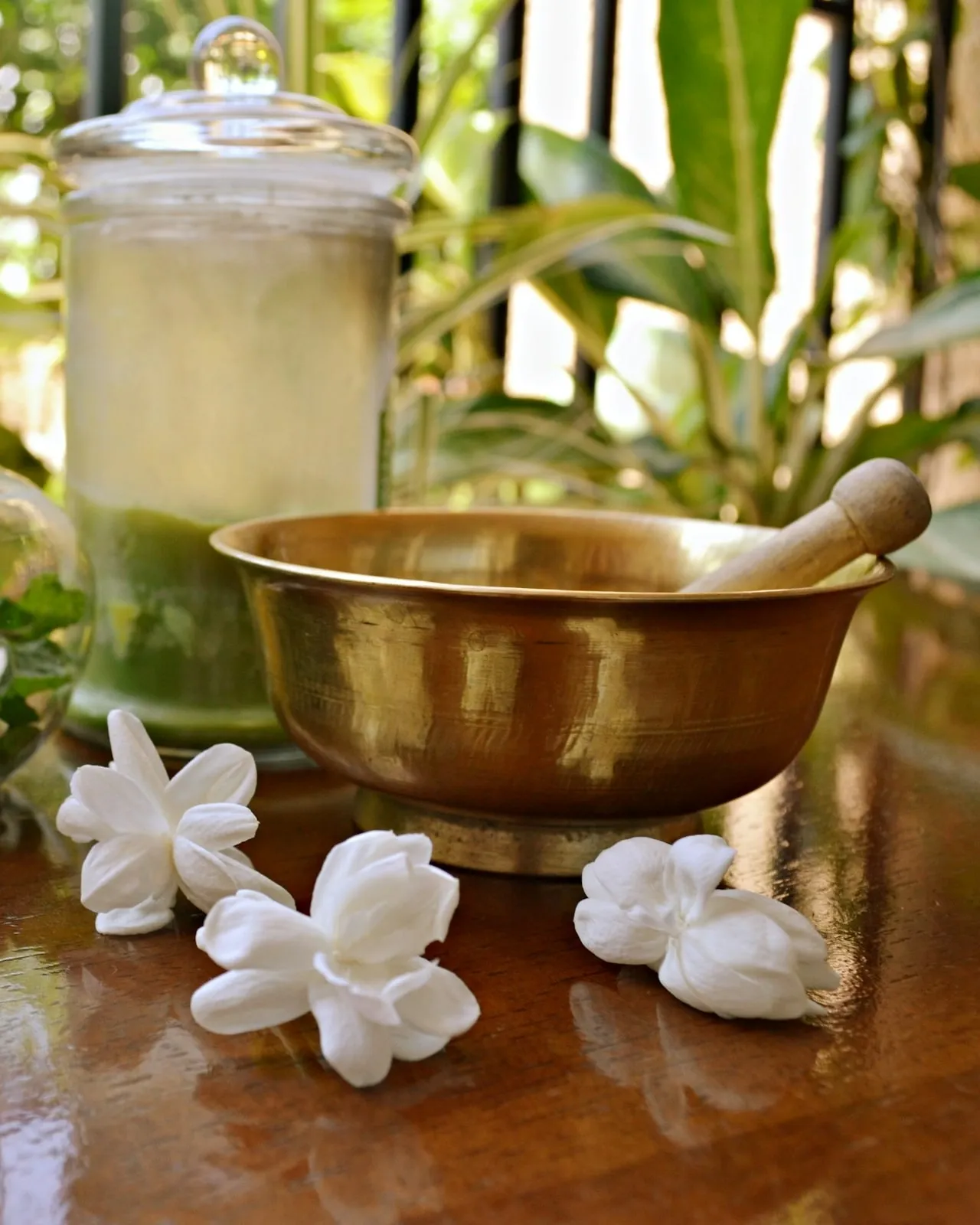a brass bowl and pestle beside a jar of herbs, with jasmine flowers on a wooden table