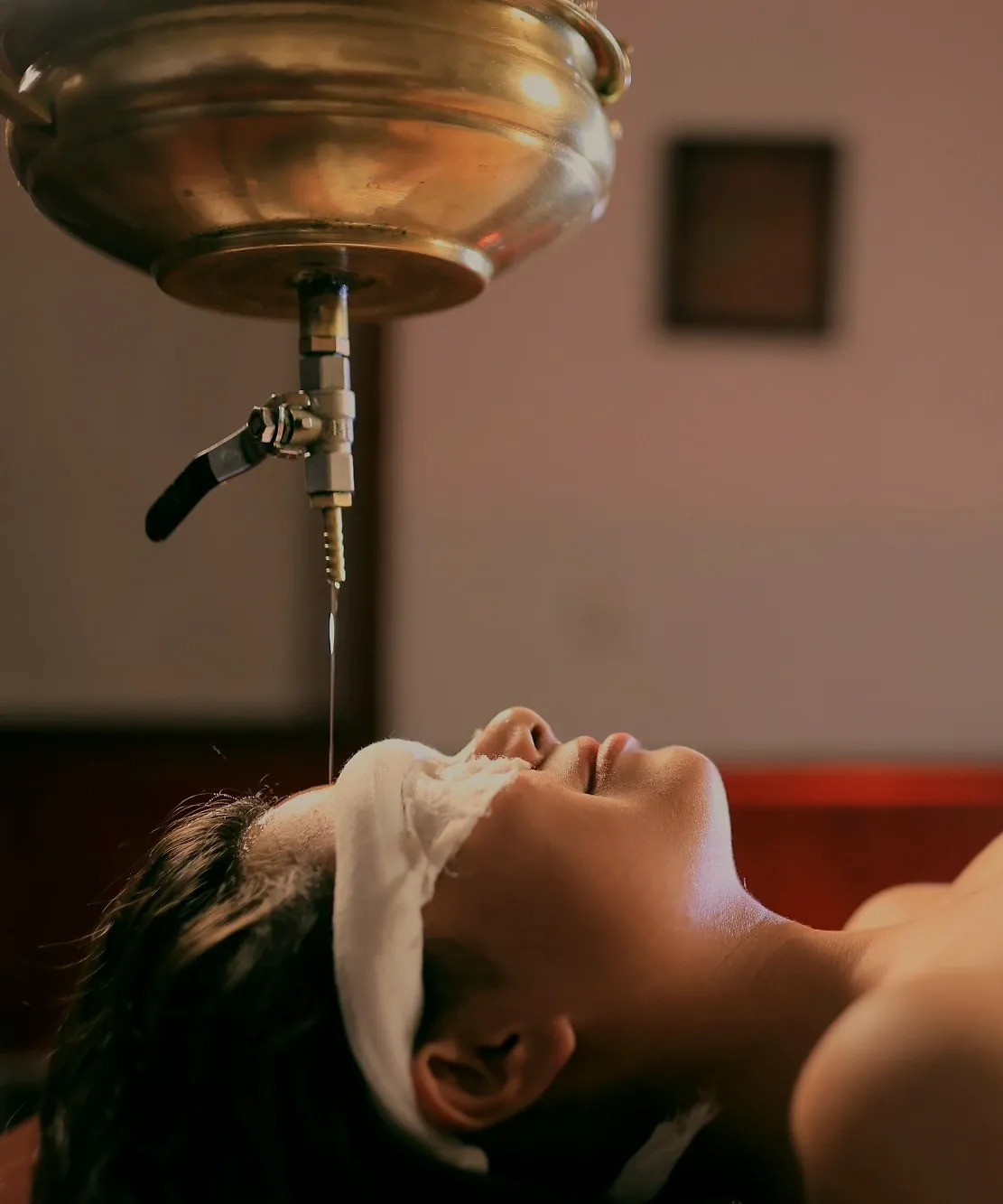 warm oil falling in a thin stream from a brass vessel onto a resting guest’s forehead